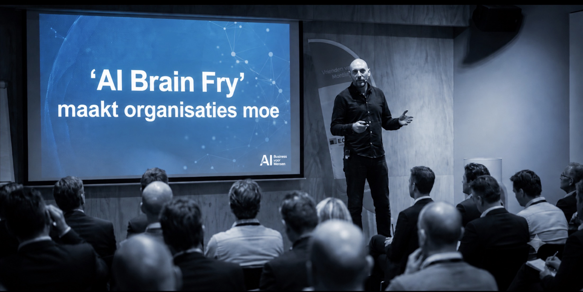 Denis Doeland spreekt voor een zaal met publiek, naast een scherm met de tekst 'AI Brain Fry' maakt organisaties moe